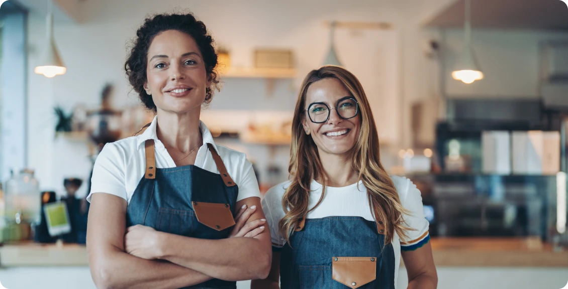 Two women in aprons smiling in café.